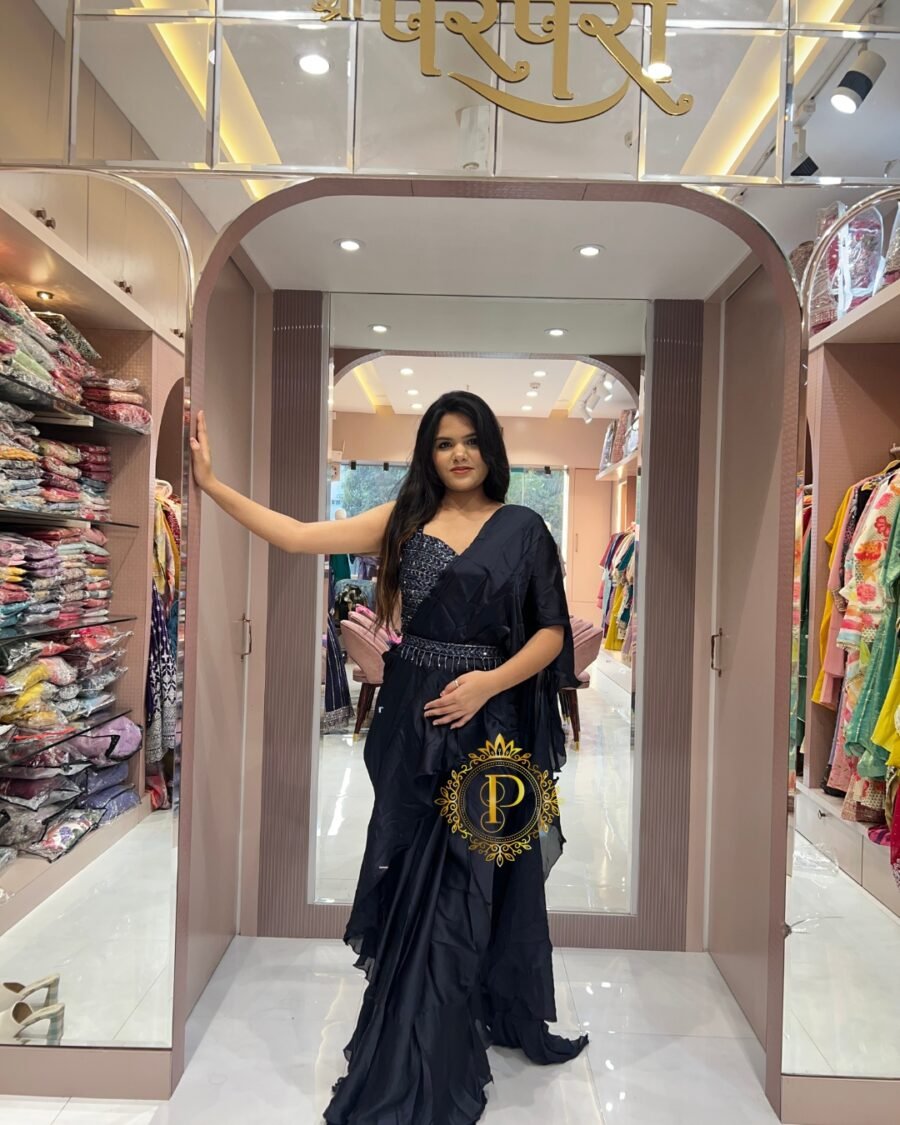 A woman poses inside a luxury ethnic wear boutique leaning against a pillar, wearing a navy blue frill saree and a hand-worked blouse.