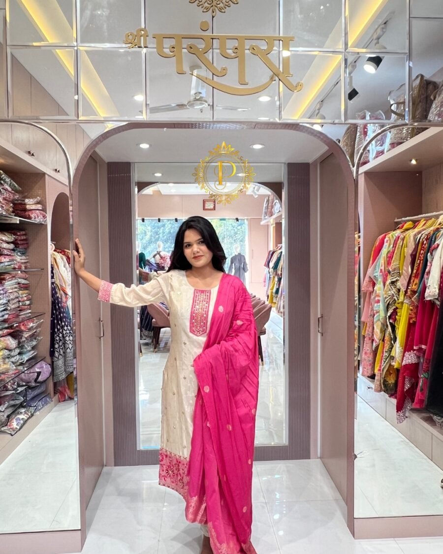 A woman standing in a doorway of a boutique wearing a shimmering ivory kurta and a bright pink dupatta with golden borders.