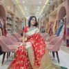 A woman sitting on a pink velvet chair in a saree shop wearing a red Banarasi print embroidery saree with green borders and gold embroidery.