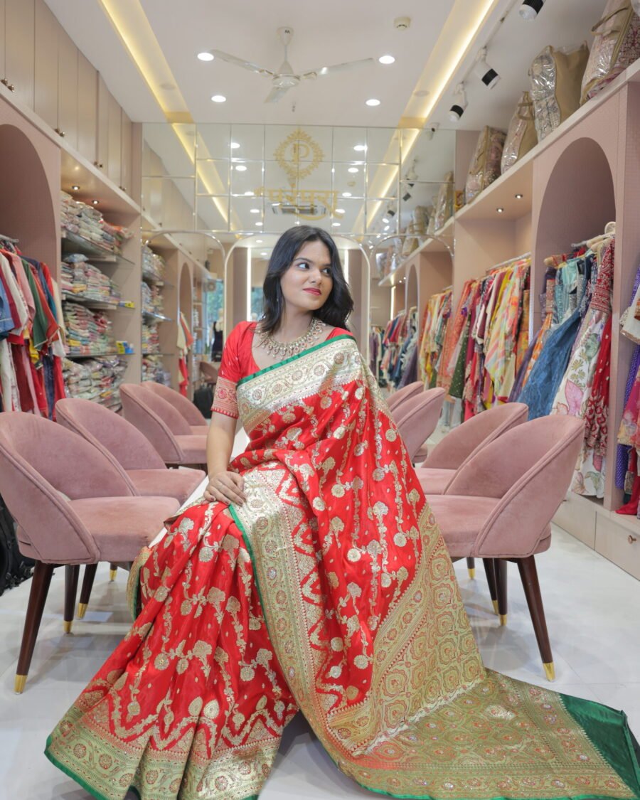 A woman sitting on a pink velvet chair in a saree shop wearing a red Banarasi print embroidery saree with green borders and gold embroidery.