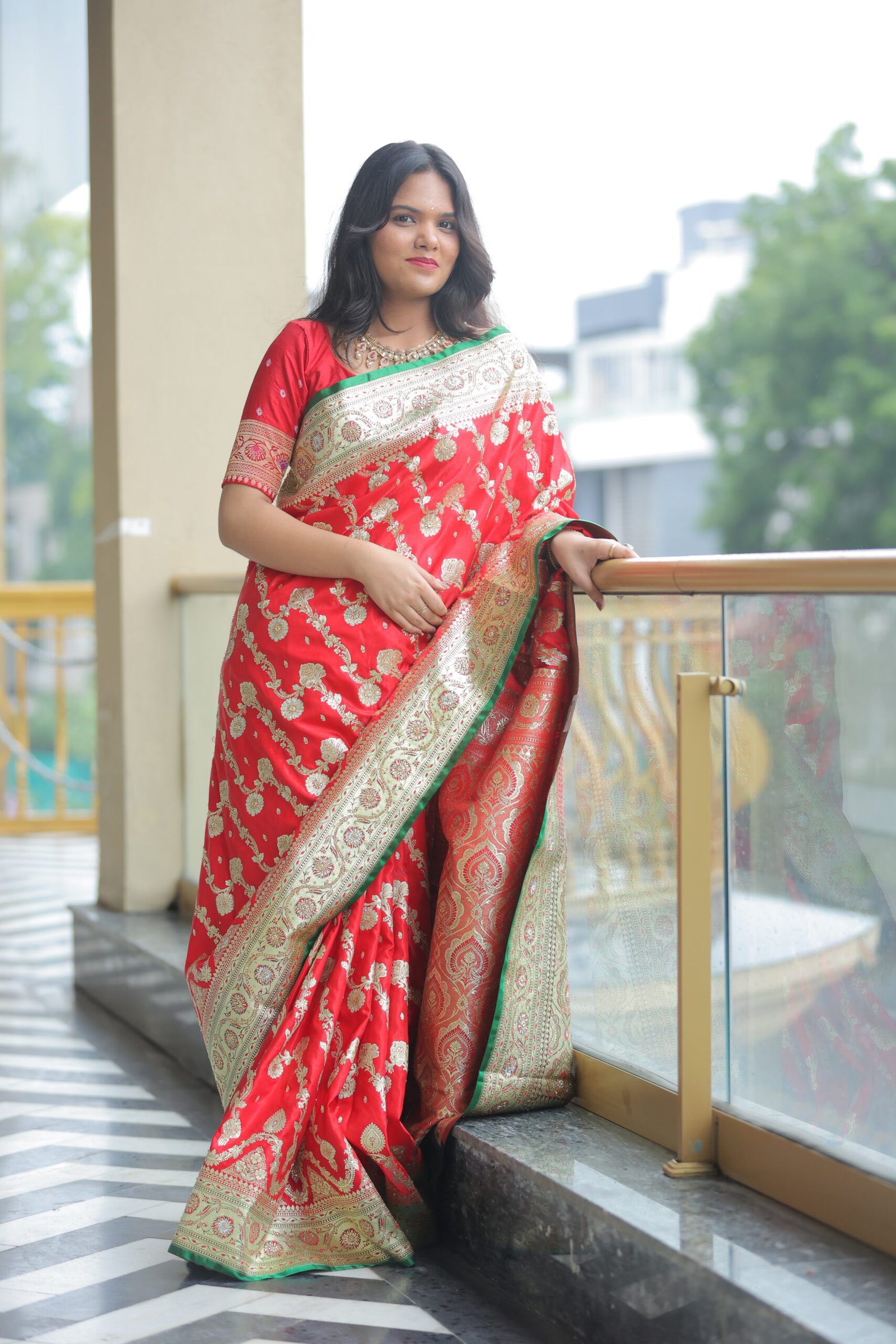 A woman standing on a balcony with a glass railing, wearing a red Banarasi saree with a heavy gold border and green edging.