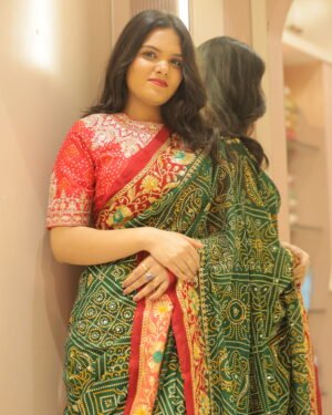 A woman standing against a neutral wall and mirror, wearing a green and red Signature Ethnic Thread Work Saree with traditional tie-dye patterns.