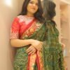 A woman standing against a neutral wall and mirror, wearing a green and red Signature Ethnic Thread Work Saree with traditional tie-dye patterns.