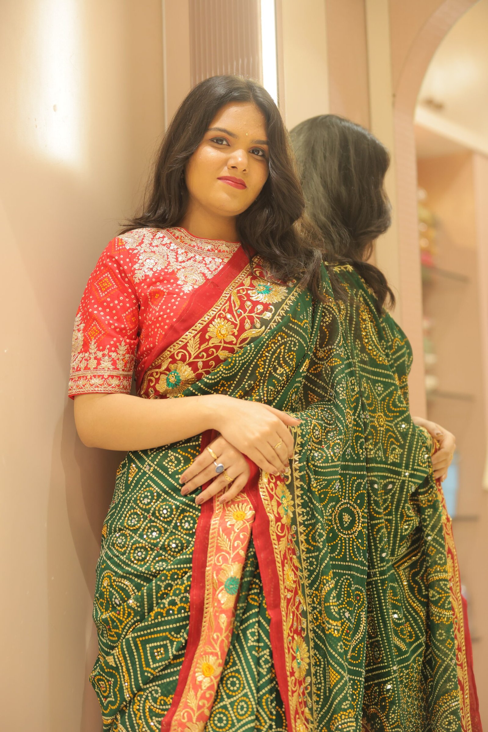 A woman standing against a neutral wall and mirror, wearing a green and red Signature Ethnic Thread Work Saree with traditional tie-dye patterns.