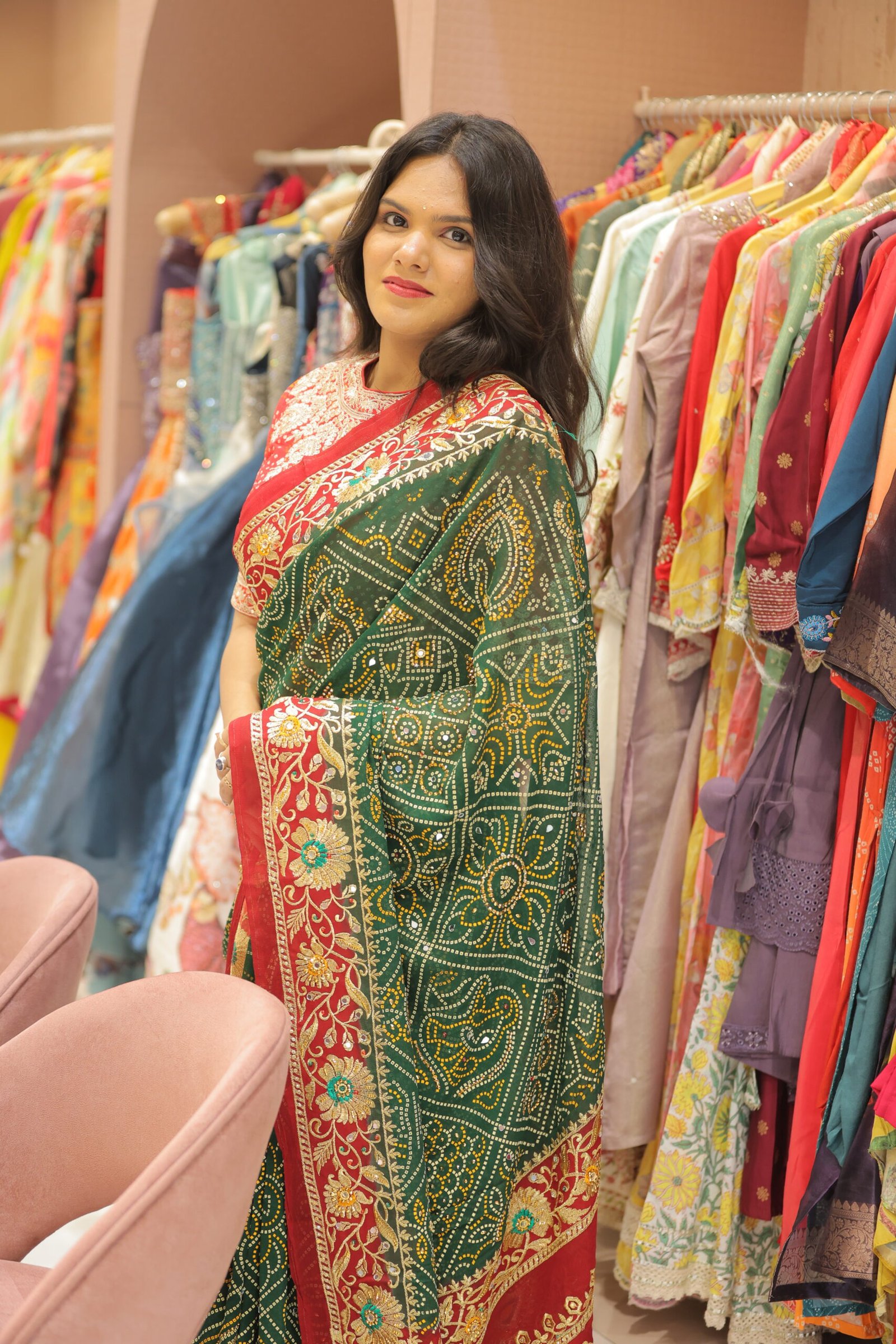 A woman posing in a green and red Bandhani saree surrounded by racks of colorful ethnic clothing.