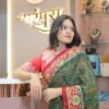 A woman leaning on a boutique counter wearing a green and red embroidered saree with a jewelry display in the background.