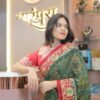A woman leaning on a boutique counter wearing a green and red embroidered saree with a jewelry display in the background.