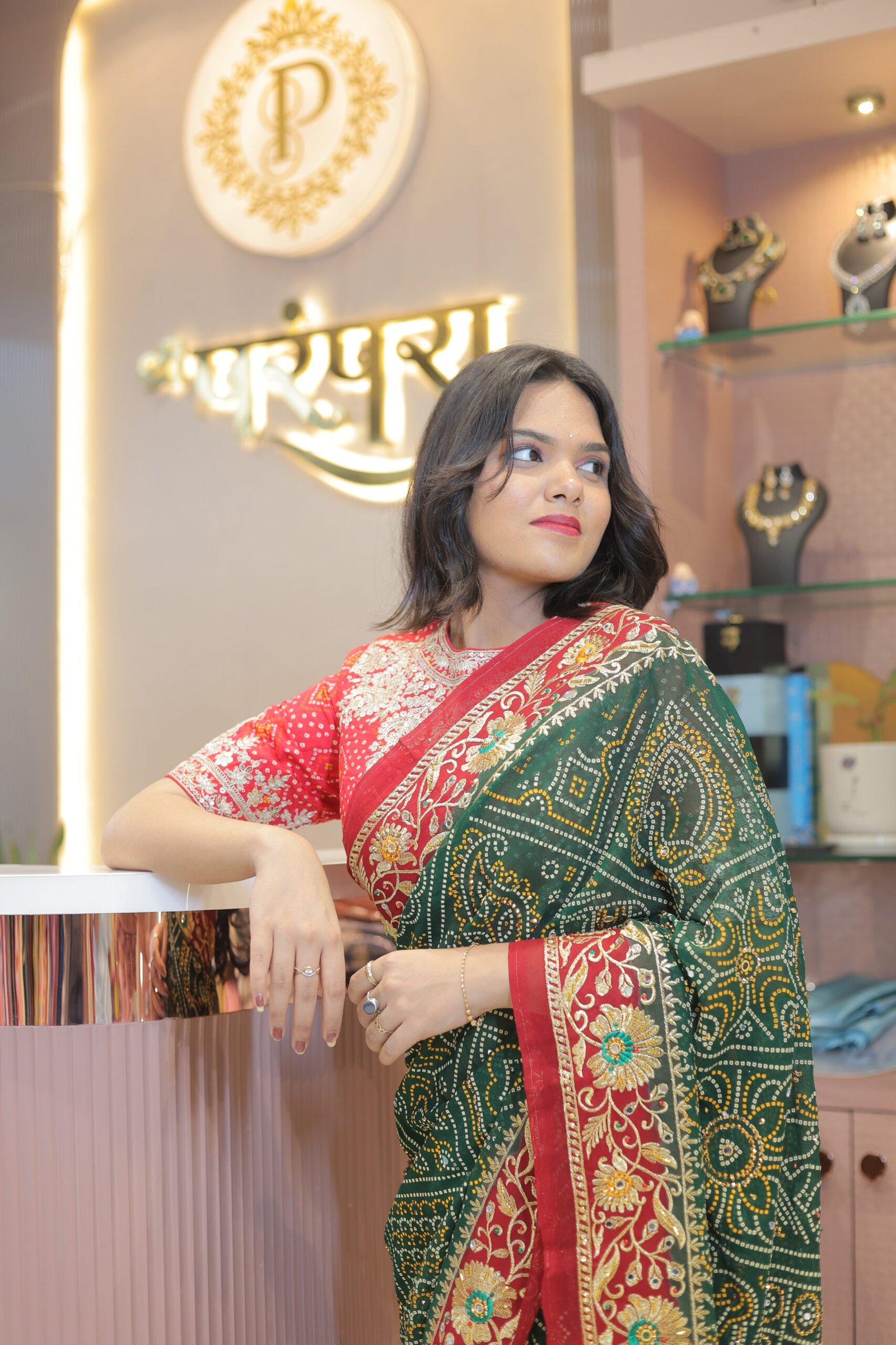 A woman leaning on a boutique counter wearing a green and red embroidered saree with a jewelry display in the background.